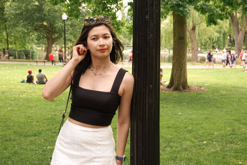 A woman leaning on a black lamppost, in a park, holding onto her ear. She is wearing a black top and white skirt and has sunglasses on her head.