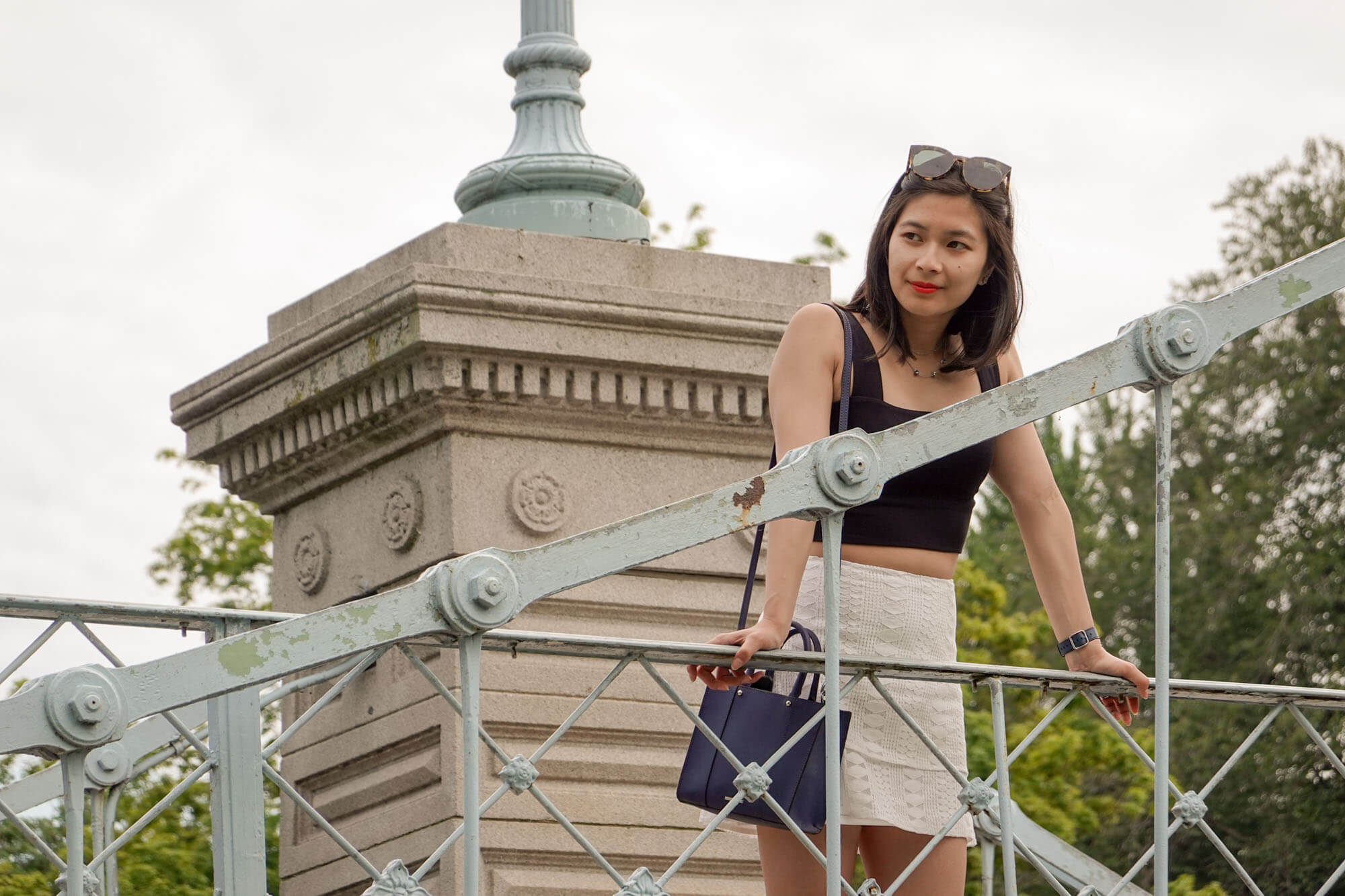 A woman leaning lightly on the railing of a bridge with her palms. She is looking into the distance and smiling slightly