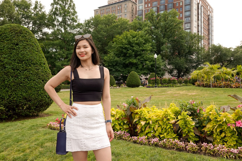 A woman standing with a green and flowery garden beind her. She has a hand on her hip.