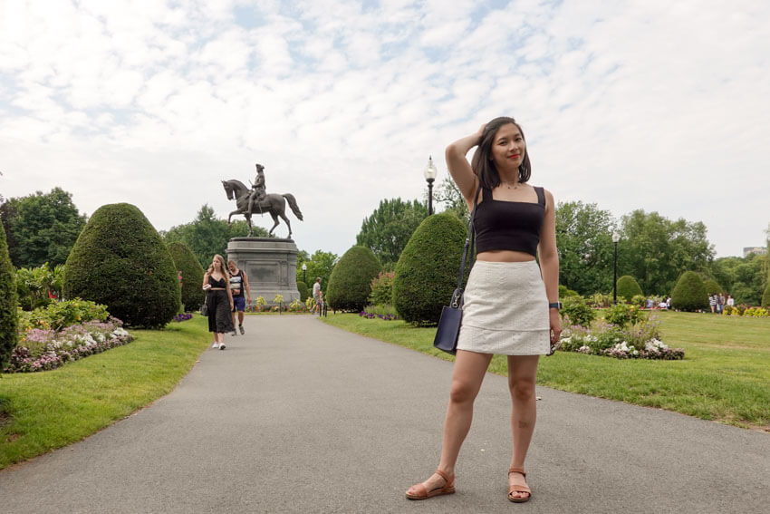 A woman standing on a pathway in a park with a lot of grass and shrubs. She has a hand on the side of her head, running it through her hair