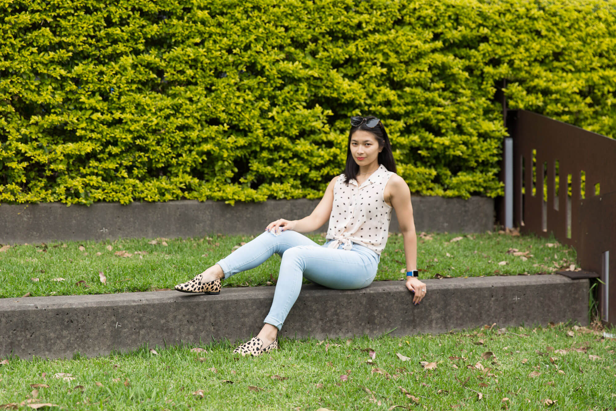 A woman sitting on the concrete step of a grassy park. Her legs are bent and slightly outstretched and she is leaning back using one of her hands as support. She is wearing light blue jeans, a light coloured top and shoes, which have patterns on them. She has sunglasses on top of her head