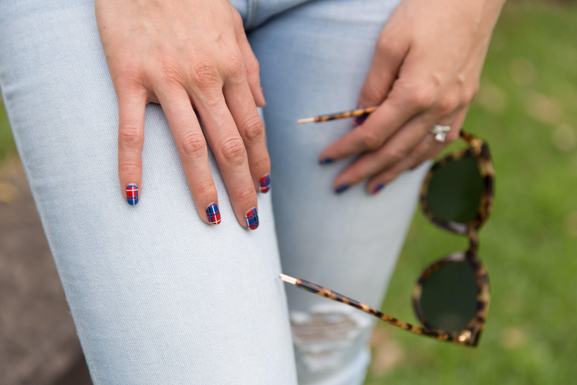 A closeup shot of a woman’s hand resting on her thigh. She has a plaid print on her fingernails and is wearing light coloured jeans. In her opposite hand she is holding a pair of glasses