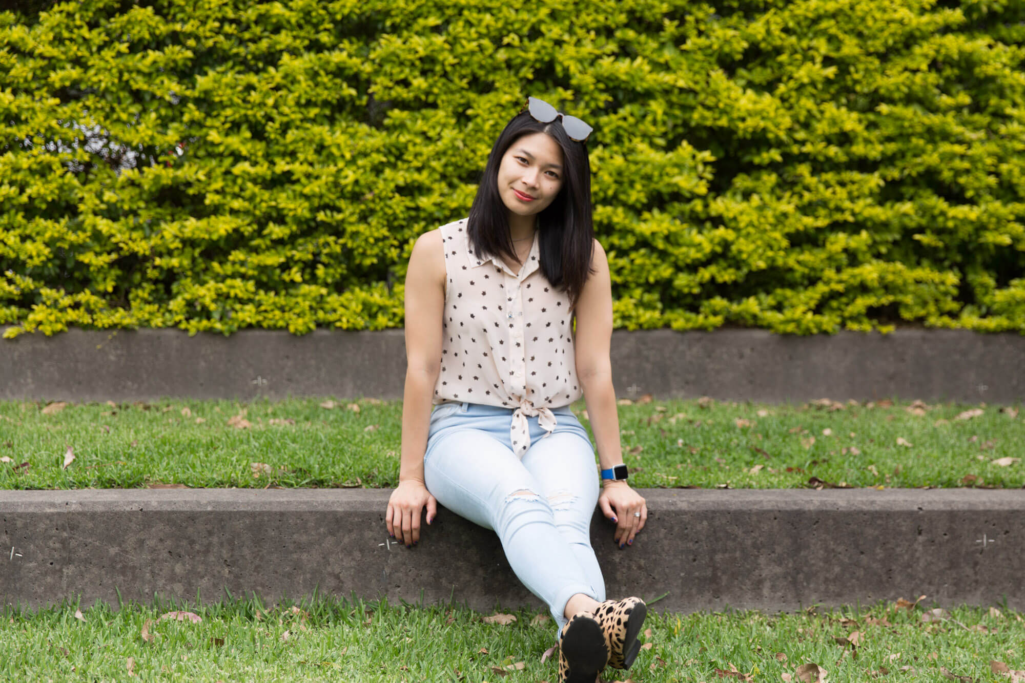 A woman sitting on a concrete step of a grassy area. She has her legs outstretched towards the camera with both hands at her sides for support