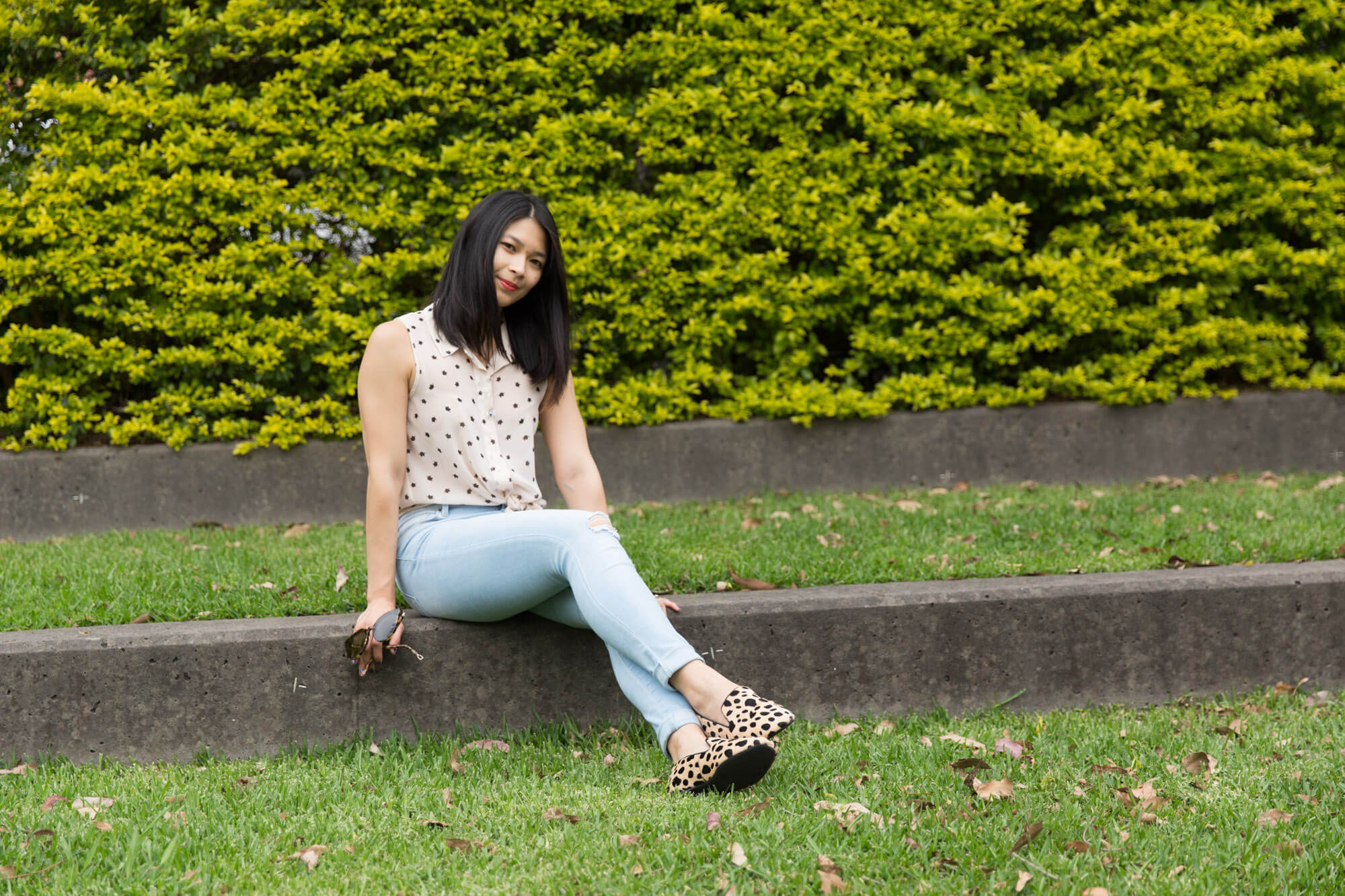A woman sitting on a concrete step of a grassy area. She has her legs outstretched towards the camera, slightly at an angle, with both hands at her sides for support. In one hand she is also holding a pair of glasses