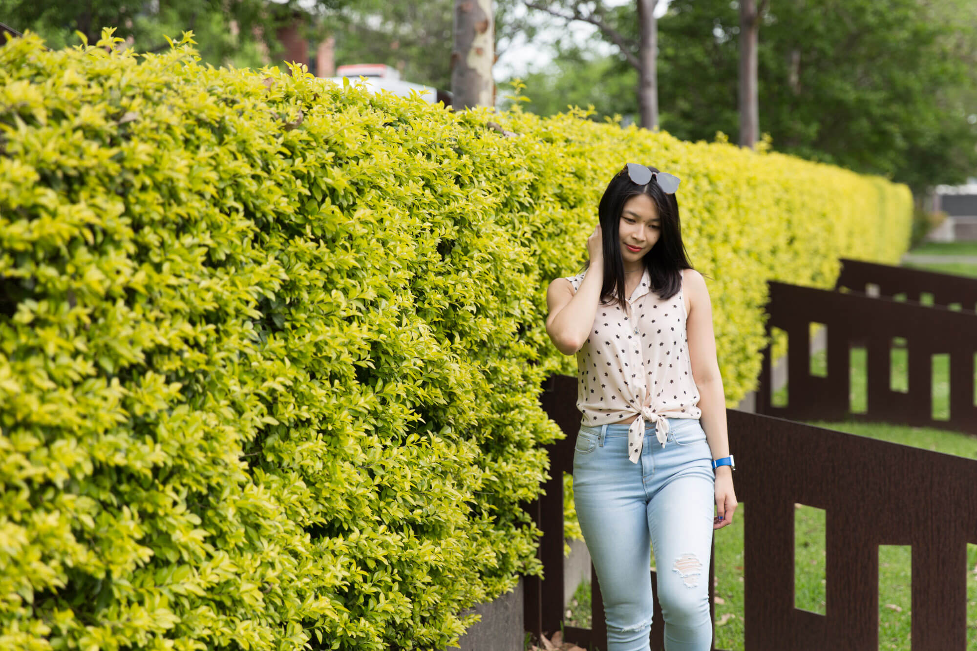 A woman standing by a bright yellow-green hedge. She has one hand by her side and the opposite palm resting on the side of her head. Her gaze is slightly down and away from the camera