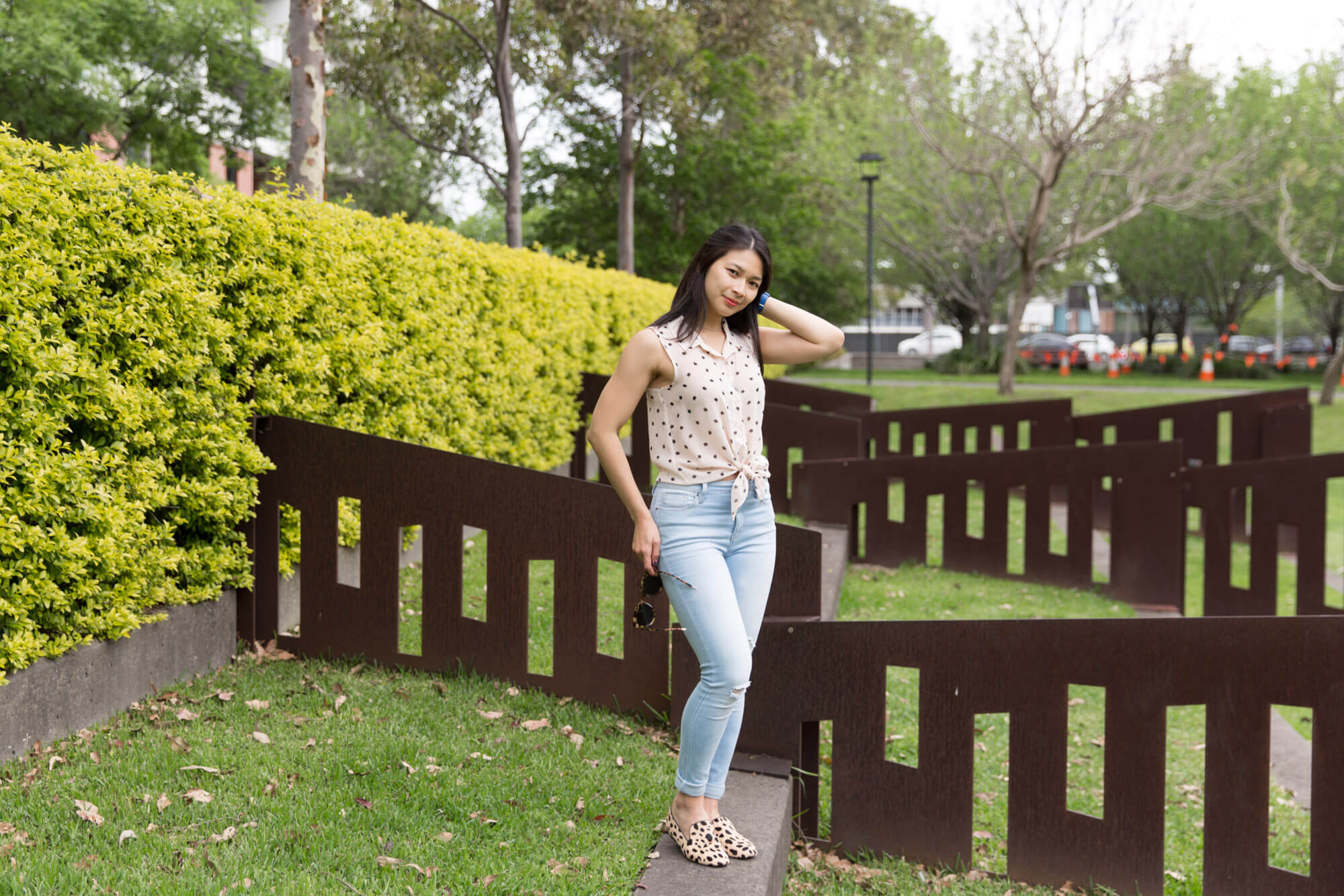 A woman standing in a park with short, dark brown angled metal fences behind her. Her feet are close together but one knee is slightly bent. She is looking at the camera and has one hand placed behind her head, and the other at her hip while holding a pair of sunglasses