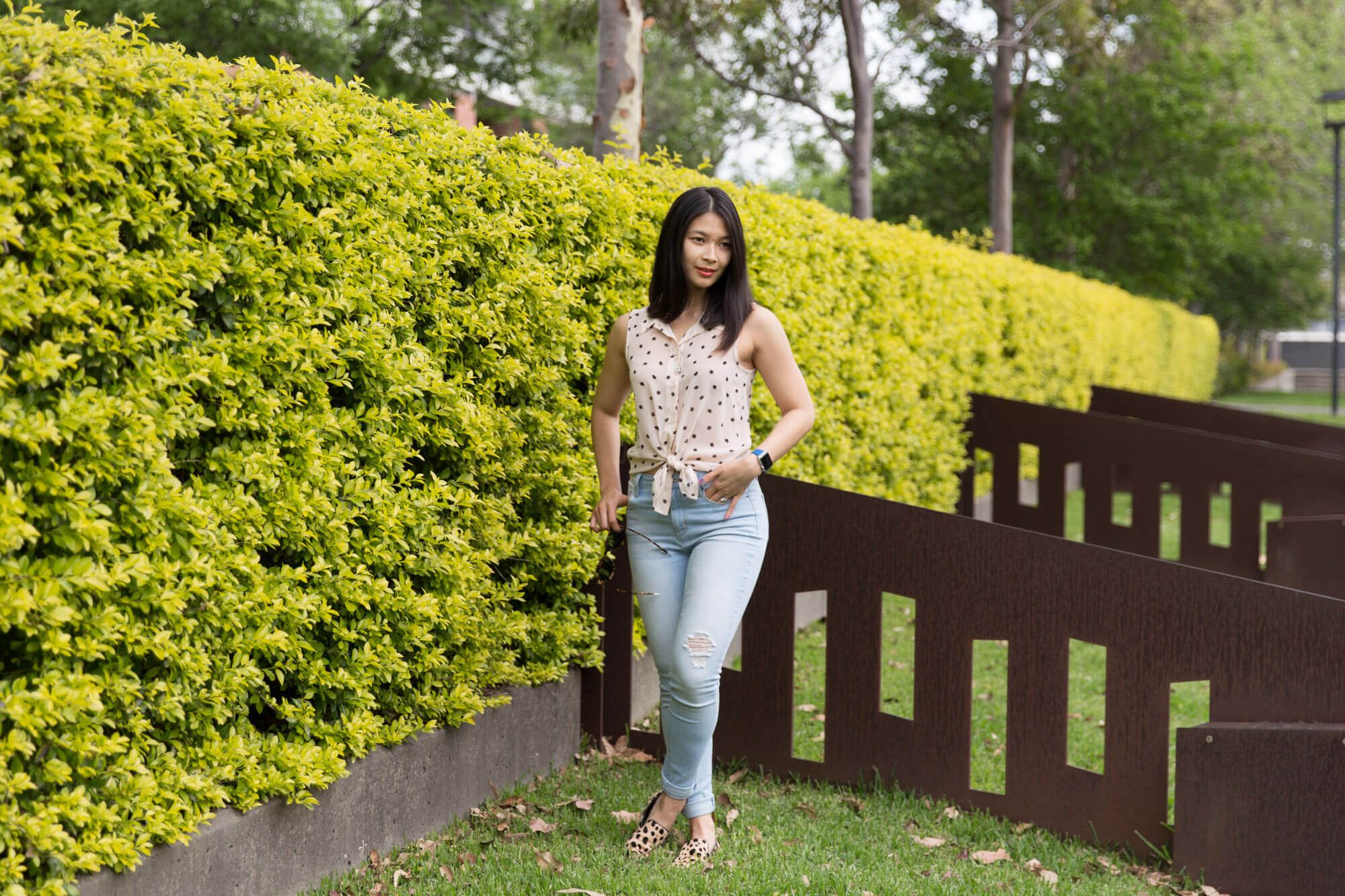 A woman standing in a park. Her feet are crossed at the ankles, and both hands are casually in her jean pockets.