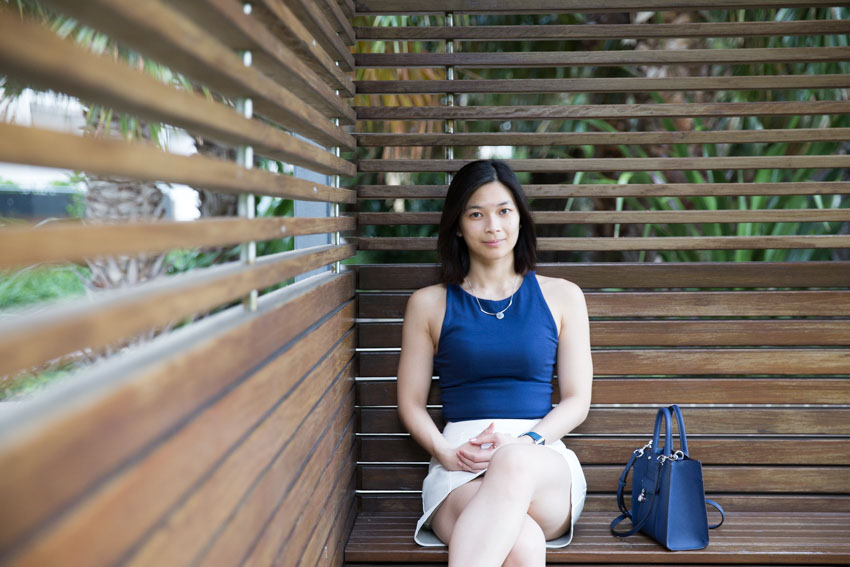 A woman with short dark hair sitting in a cabana facing the camera. She is wearing a blue sleeveless top and has a small square-shaped blue bag next to her.