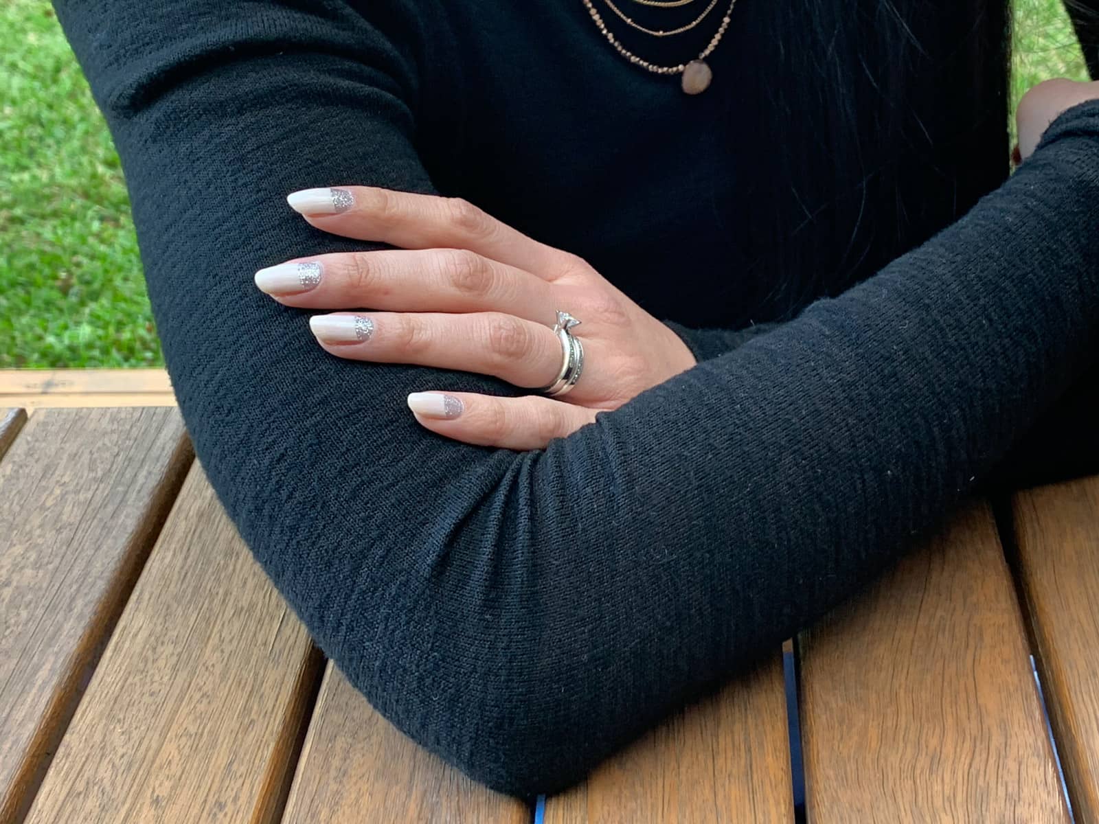 A close-up of a woman’s arms, folded. She has nail art on her fingernails – silver glitter near the cuticles, and the remainder a light beige colour. She is wearing a silver wedding and engagement ring on her fourth finger.