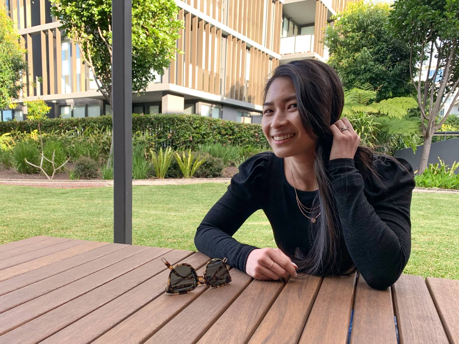 A woman wearing a long-sleeved black top, sitting at a wooden table, with both elbows on the table. She is holding her long hair away from her face with one hand. The other hand rests on the table alongside a pair of tortoiseshell sunglasses.