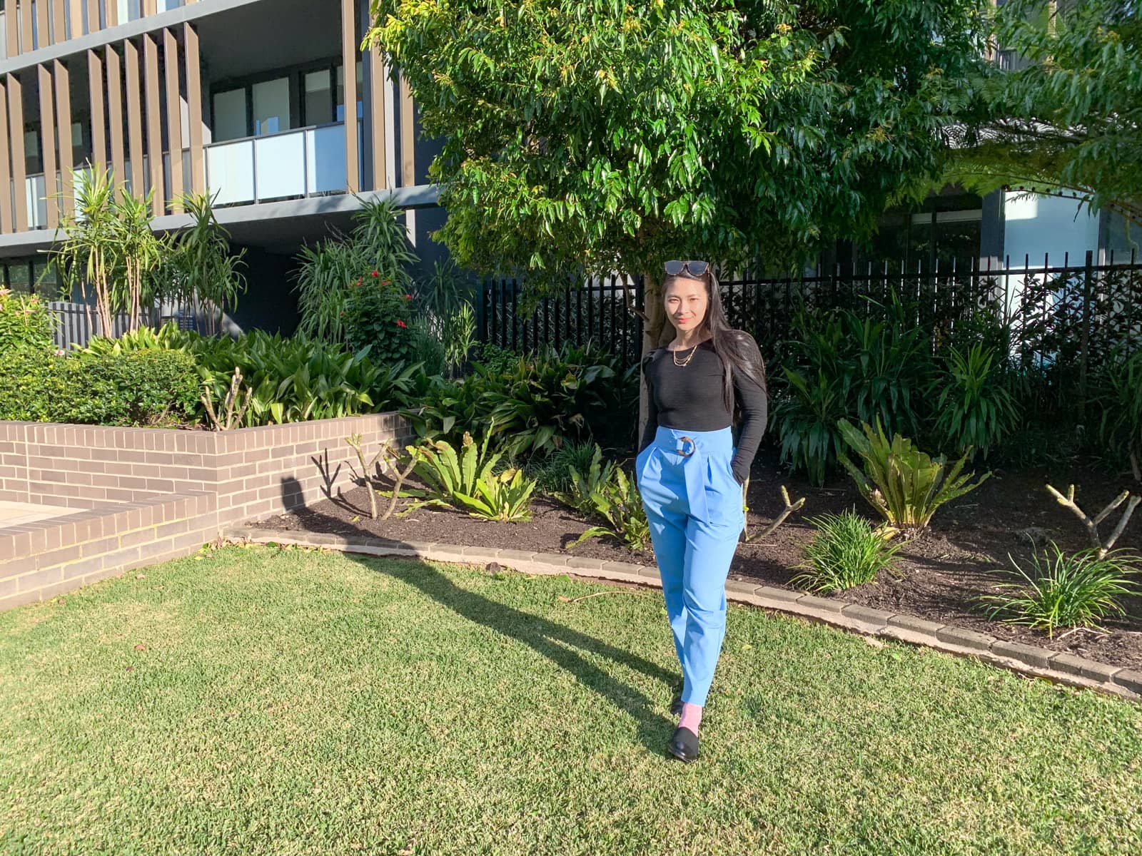 A woman with long dark hair, wearing a long-sleeved black top and sky blue pants. She has pink socks and is wearing black loafers. Her hands are in her pockets and she is leaning back on one foot with her other foot in front. She is standing on grass in a common area with some high-rise apartments behind her.