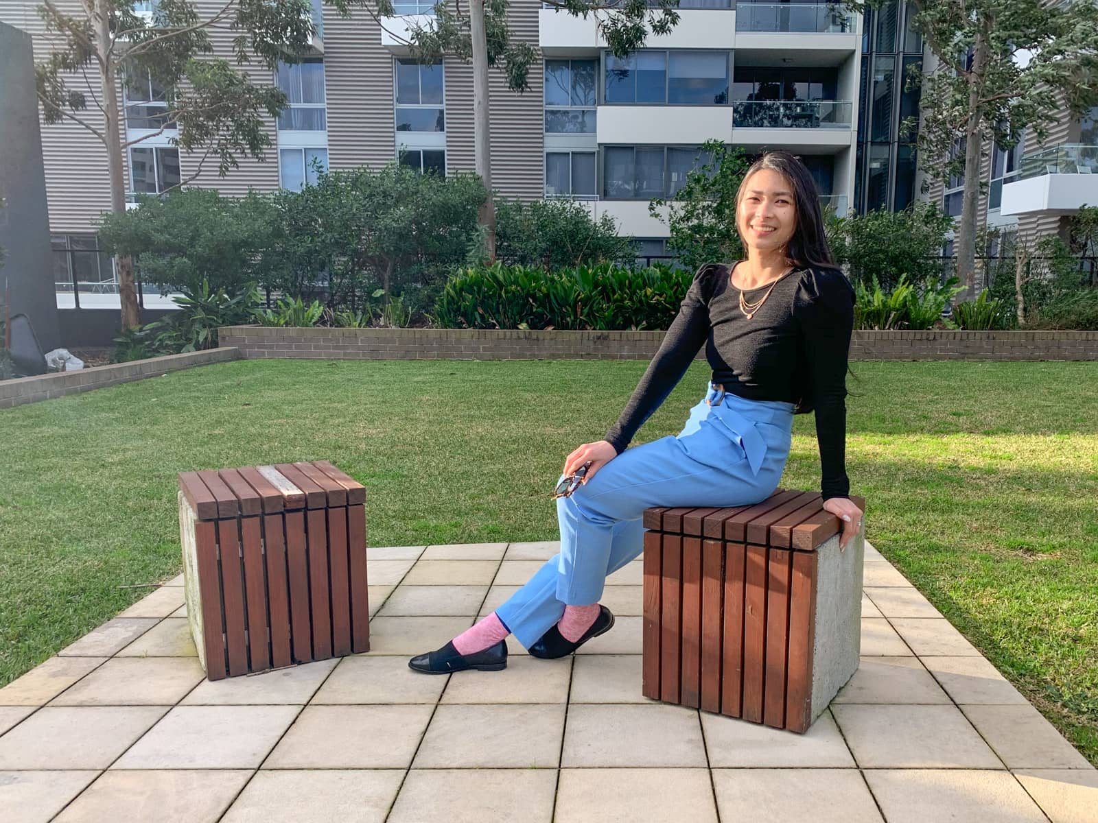 A woman sitting on a concrete cube-shaped block topped with wooden planks, made to function as a stool. She is leaning back on her hand, which is resting on an edge of the block. One of her legs is outstretched with her foot on the ground, and the other leg is bent. She is wearing a long-sleeved black top with sky blue pants and is holding a pair of sunglasses in her free hand.