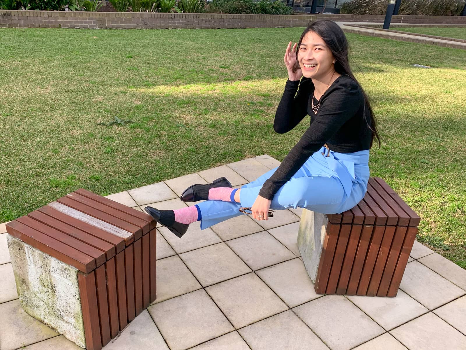 A woman sitting on a concrete cube-shaped block topped with wooden planks, made to function as a stool. She has her feet propped up on another wooden block. She is wearing a black long-sleeved top and sky blue pants, and is smiling and tucking hair behind her ear.