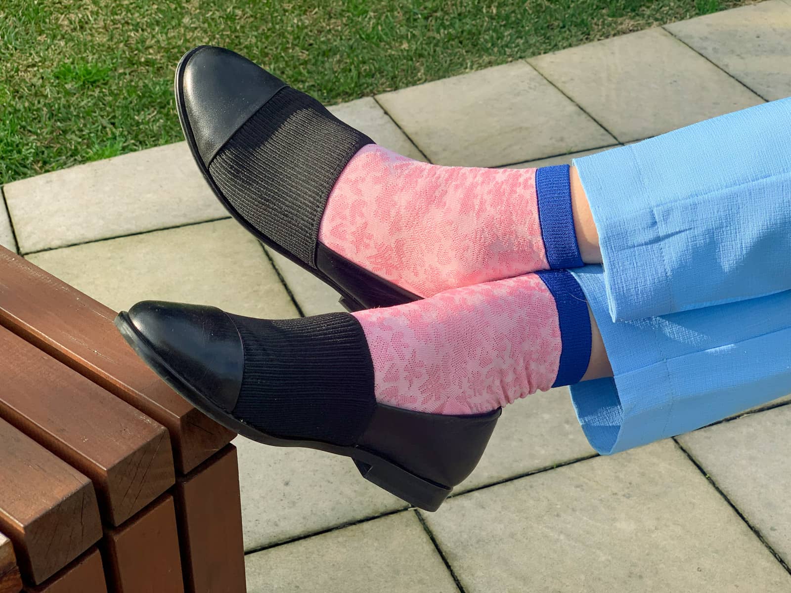 A close-up of a womanâ€™s feet wearing pink socks with a splatter texture, with blue top edges. She is wearing black loafers on her feet that have a knitted area covering the top part of the loafer