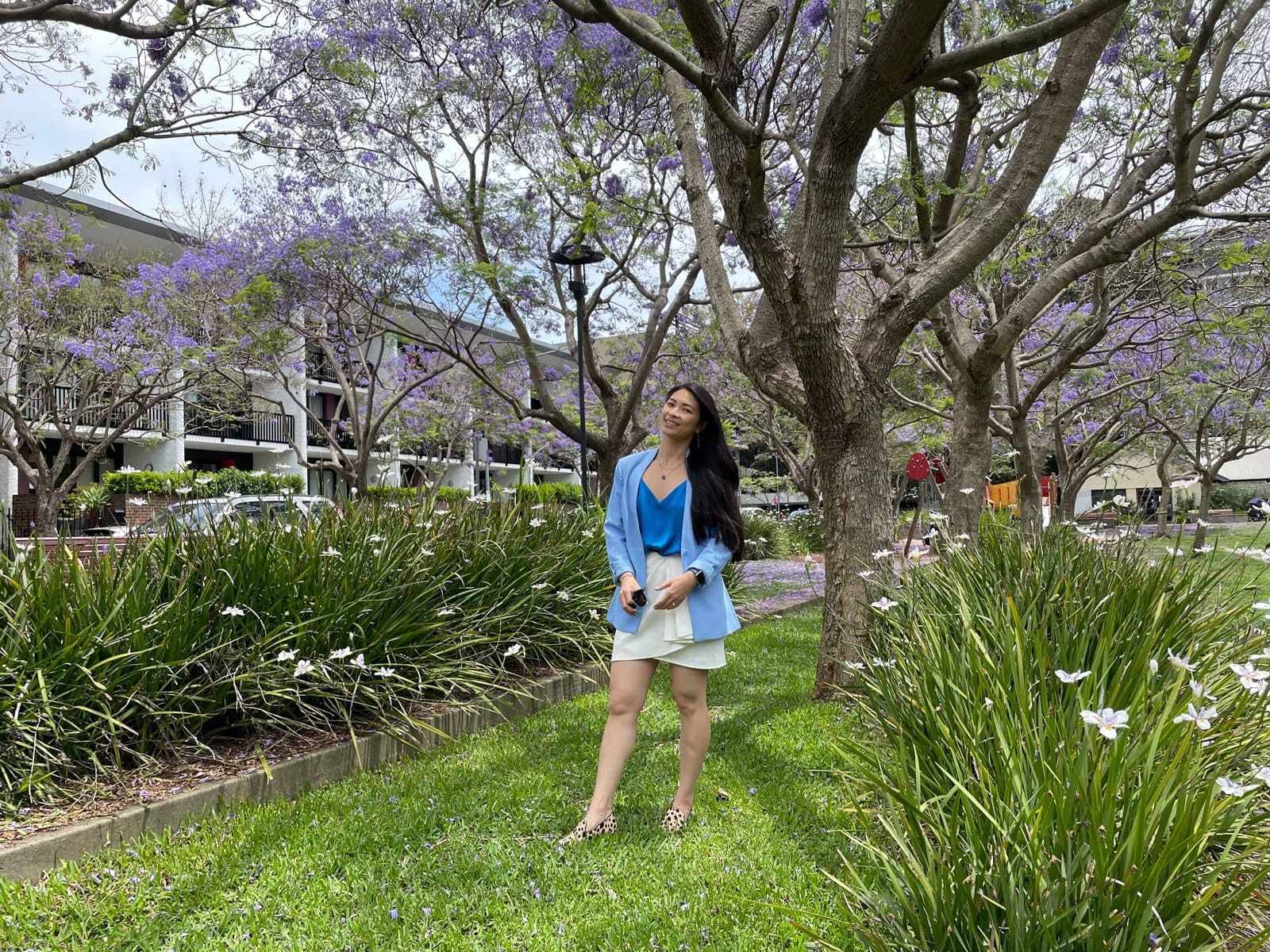 A woman with long dark hair, wearing a blue blazer over a blue top and white skirt, standing in a lush green park with jacaranda trees. She has loafers with a giraffe animal print