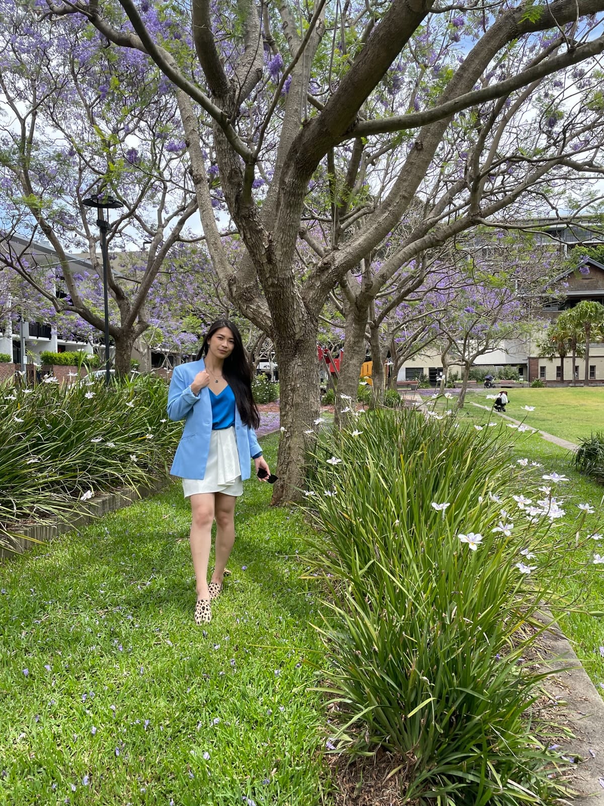 The same woman in previous photos in this post, wearing the same outfit and in the same location. She is holding sunglasses in one of her hands and standing with one leg slightly in front of the other.