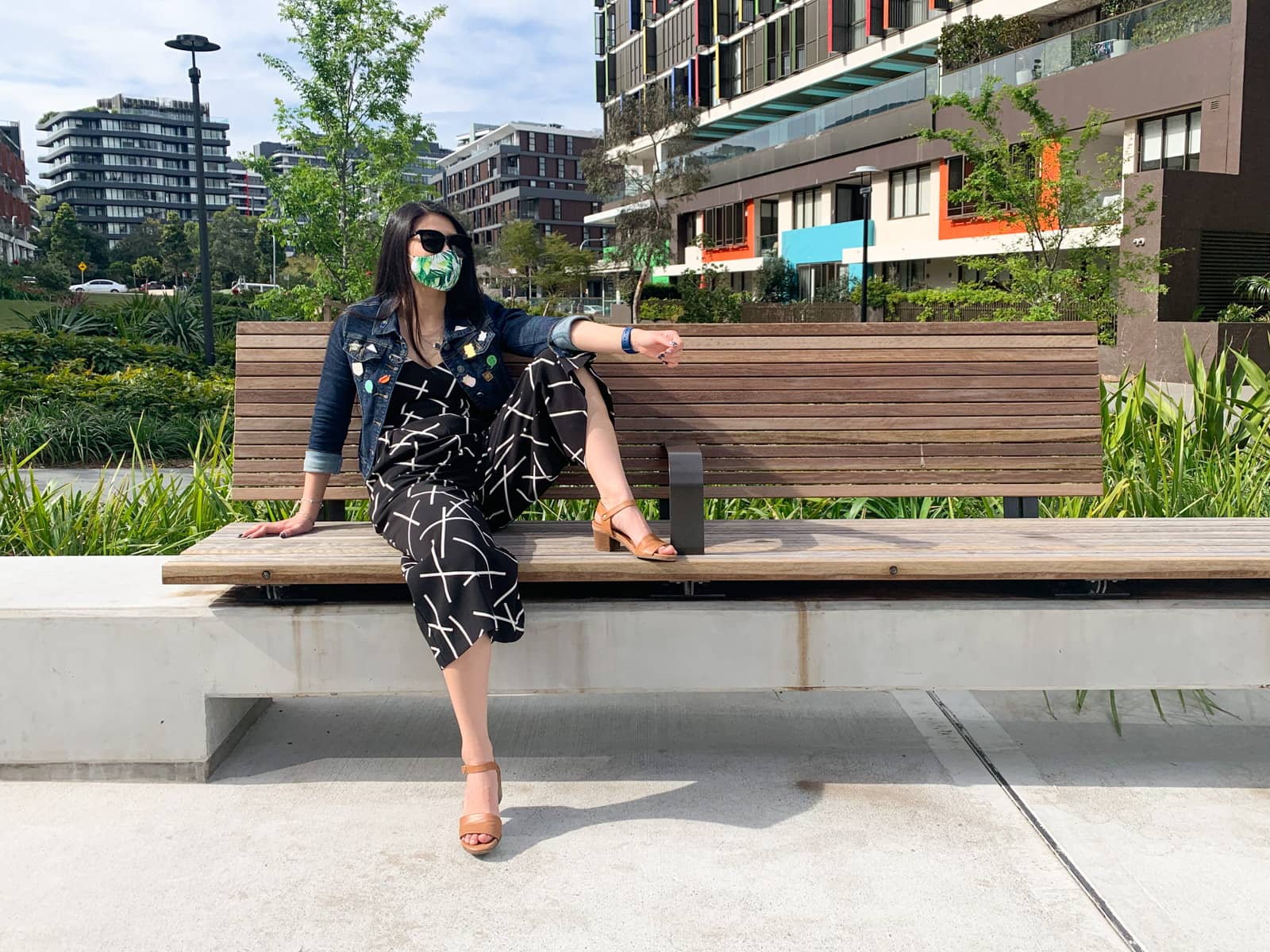 The same woman in previous photos on this page, in the same setting and wearing the same clothes. She is sitting on a wooden bench with one foot up on the bench and her knee bent. She has dark black sunglasses on.