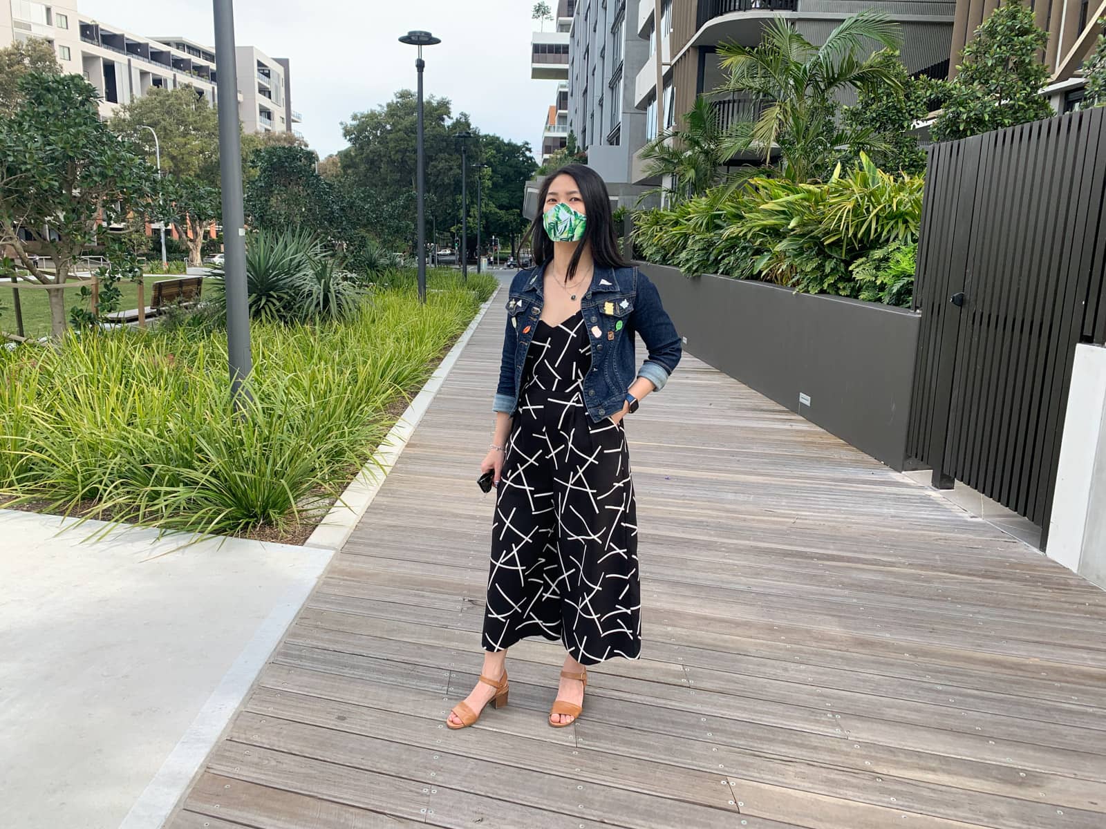 The same woman in previous photos on this page, wearing the same clothes and standing on a boardwalk with some gardens and apartments in the background. She is standing and has a hand in her pocket.