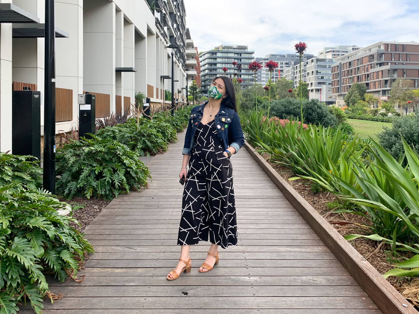 A woman with long dark hair, wearing a wide leg black jumpsuit with white geometric lines on it. She is wearing a denim jacket over the jumpsuit and has one hand in a pocket. She is wearing tan sandals, and also wearing a face mask with a leaf print. In the background is a wooden boardwalk, with apartments down one side and plants and grass on the other.