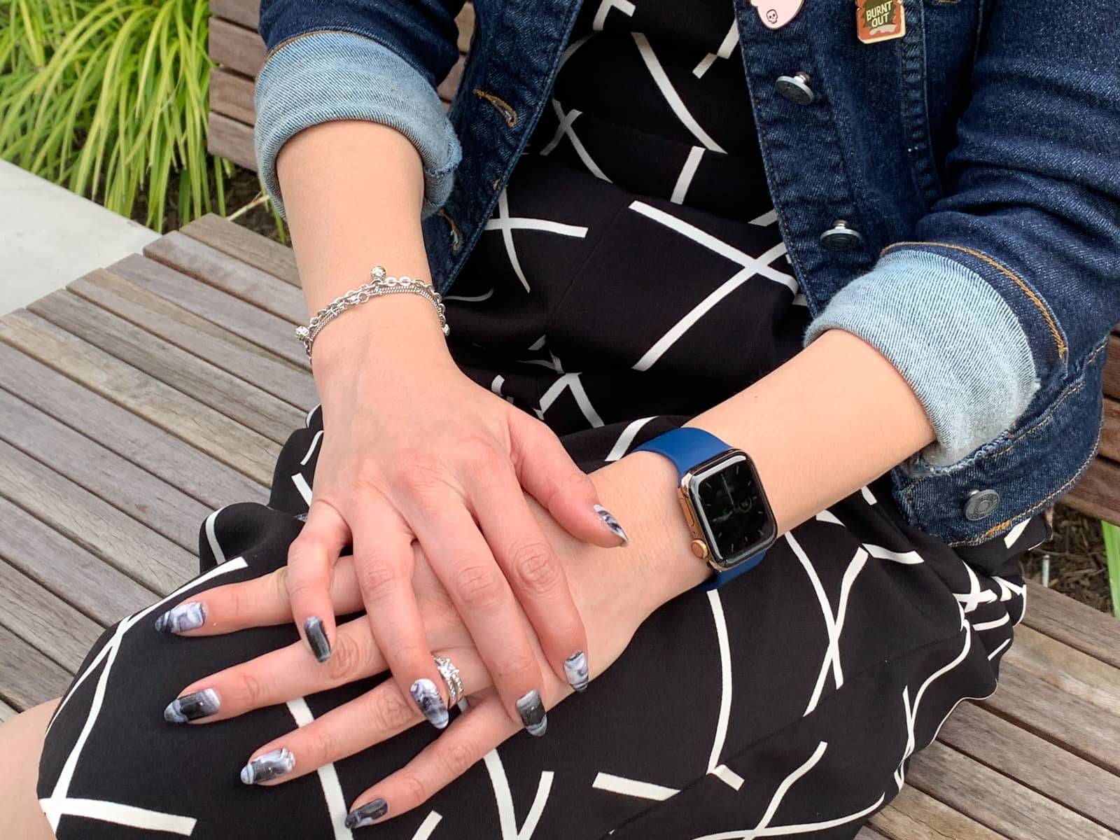 A close-up of a woman’s hands, crossed over each other, on her knee. The woman’s fingernails have black and white swirly nail art. She is wearing a blue watch on one wrist and a couple of bracelets on the other. She is wearing a black jumpsuit with white geometric lines on it.