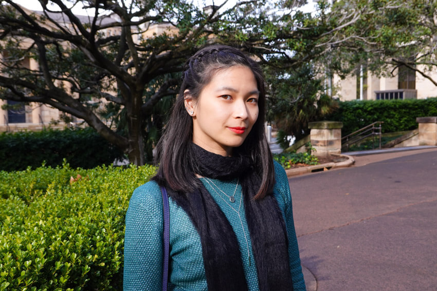 A woman with short dark hair with a braid on one side, wearing an emerald green black scarf. She has a neutral expression. She is in a park with trees in the background.