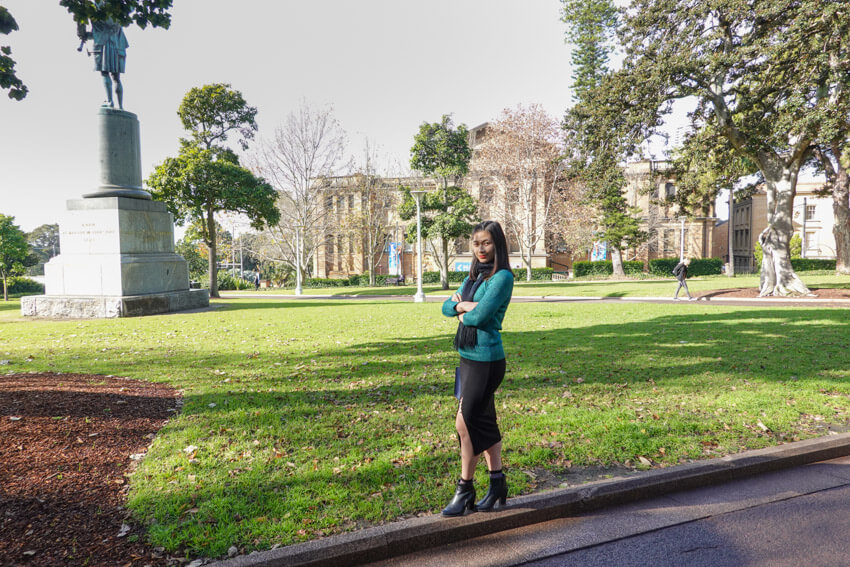 The same woman from the previous photo, wearing the same clothes and in the same setting. She has her arms folded and is standing on the raised edge on the side of a footpath.