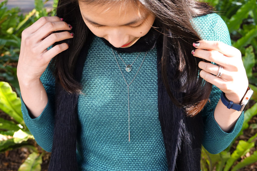 The same woman from the previous photo, wearing the same clothes. This is a closeup of her lariat necklace. Her hands hold her hair away from her chest area and she is looking down.