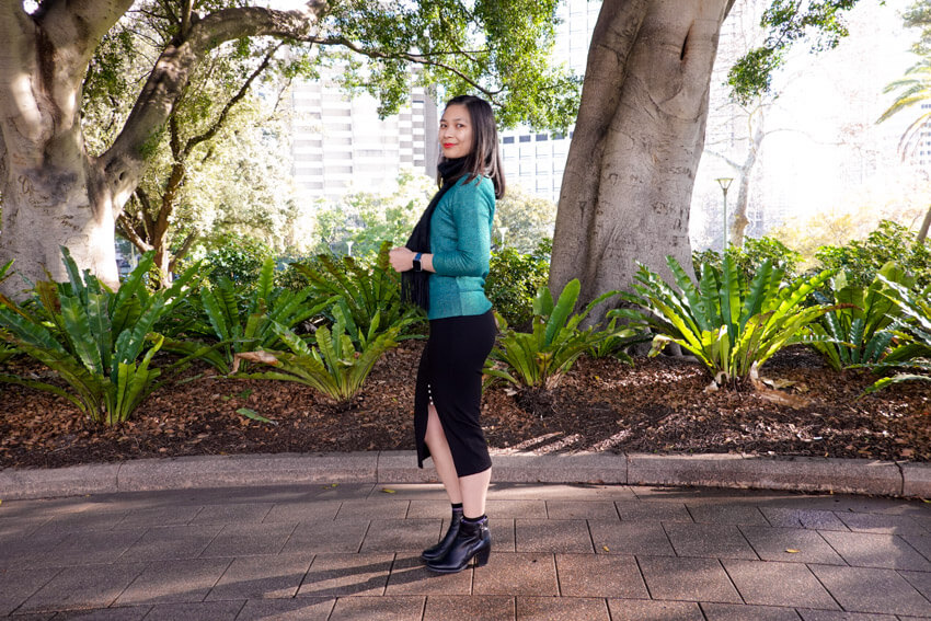 The same woman from the previous photo, wearing the same clothes and in the same setting. She is wearing a mid-calf length black skirt and black ankle boots. She is standing with her left side to the camera and her head turned towards the camera.