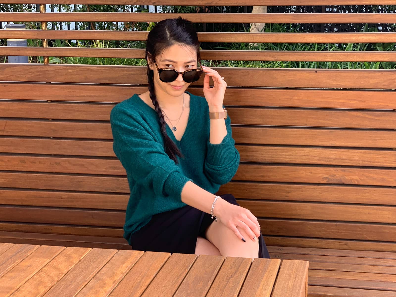 A woman with long dark hair, wearing a green sweater and black skirt, sitting in a wooden cabana, behind a wooden table. She has a braid over one of her shoulders. One of her hands rests on her knee, which is crossed over the other. Her other hand is raised to her sunglasses which are partially obscuring her face