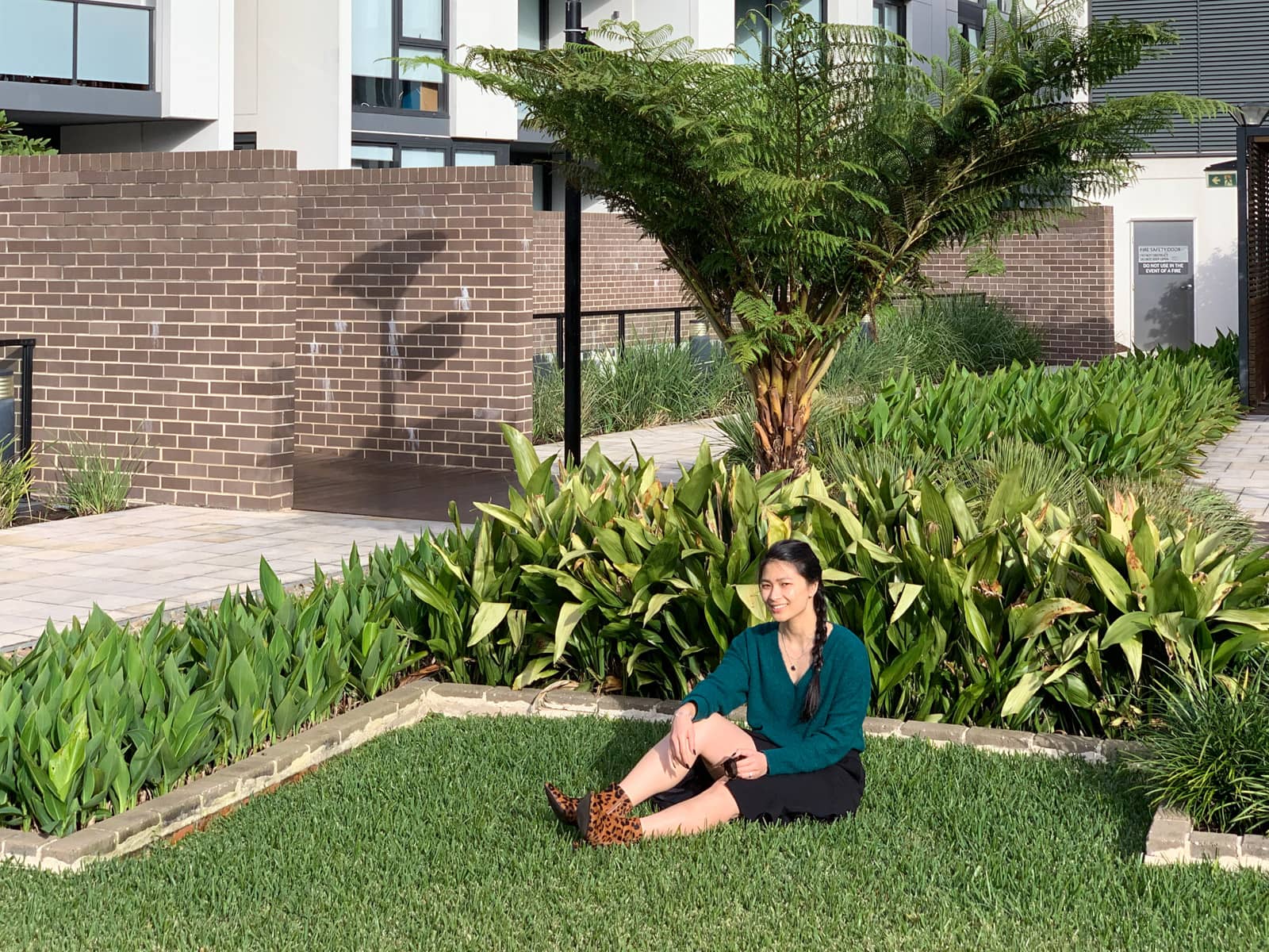 The same woman in other photos on this page, sitting down in the grass of a garden. There are some plants behind her. She is sitting with legs outstretched and one knee up