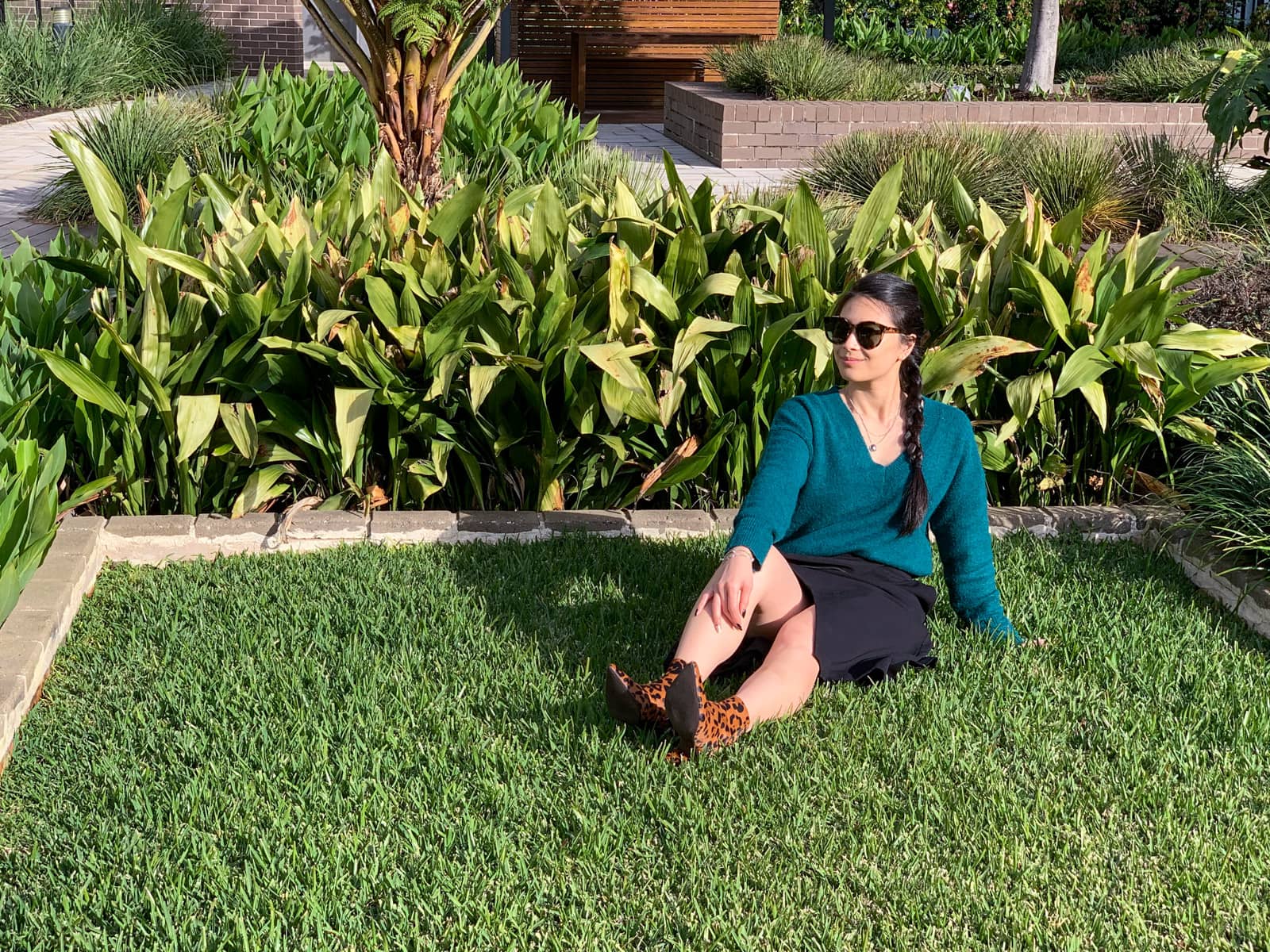 The same woman in other photos on this page, sitting down in the grass of a garden. She has sunglasses on and is looking out into the distance.