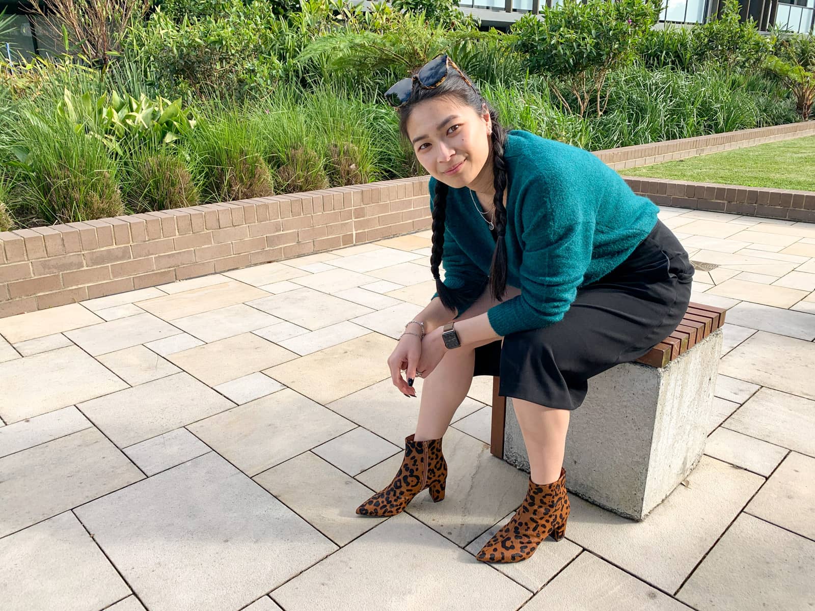 A woman dressed in a green sweater and black skirt, sitting on a concrete block topped with a wooden surface. She is resting her elbows on her thighs and looking at the camera. Her hair is in two side braids. Her shoes are brown ankle boots with black animal print. She has sunglasses on top of her head.