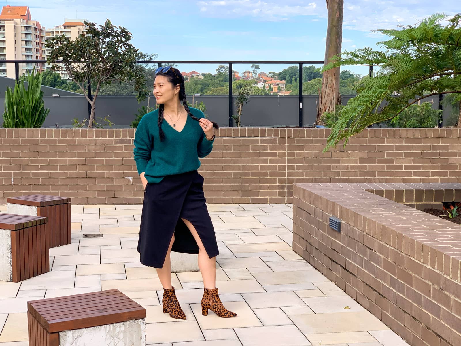 A woman with dark hair in two braids, standing in a concrete park. Some concrete cubes are fixed on the ground near her. She is lightly holding one of her two braids and looking to the left.