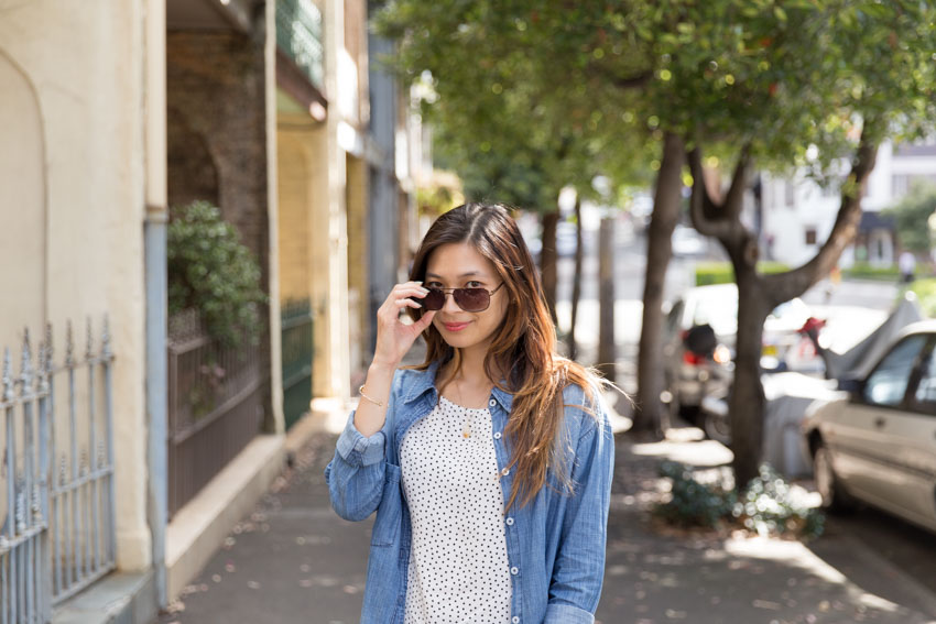 Medium shot of a woman with brown hair with blonde highlights, with her hand at my sunglasses and looking over the top of them. She is wearing a chambray shirt over a white shirt