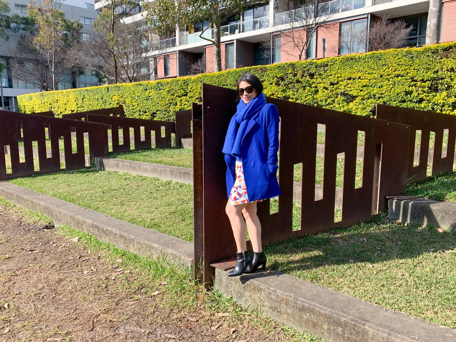 A woman in a bright blue coat and bright blue scarf standing next to a small section of fence in a park. She has sunglasses on and her hands in her coat pockets. She is wearing a colourful short skirt and black boots.