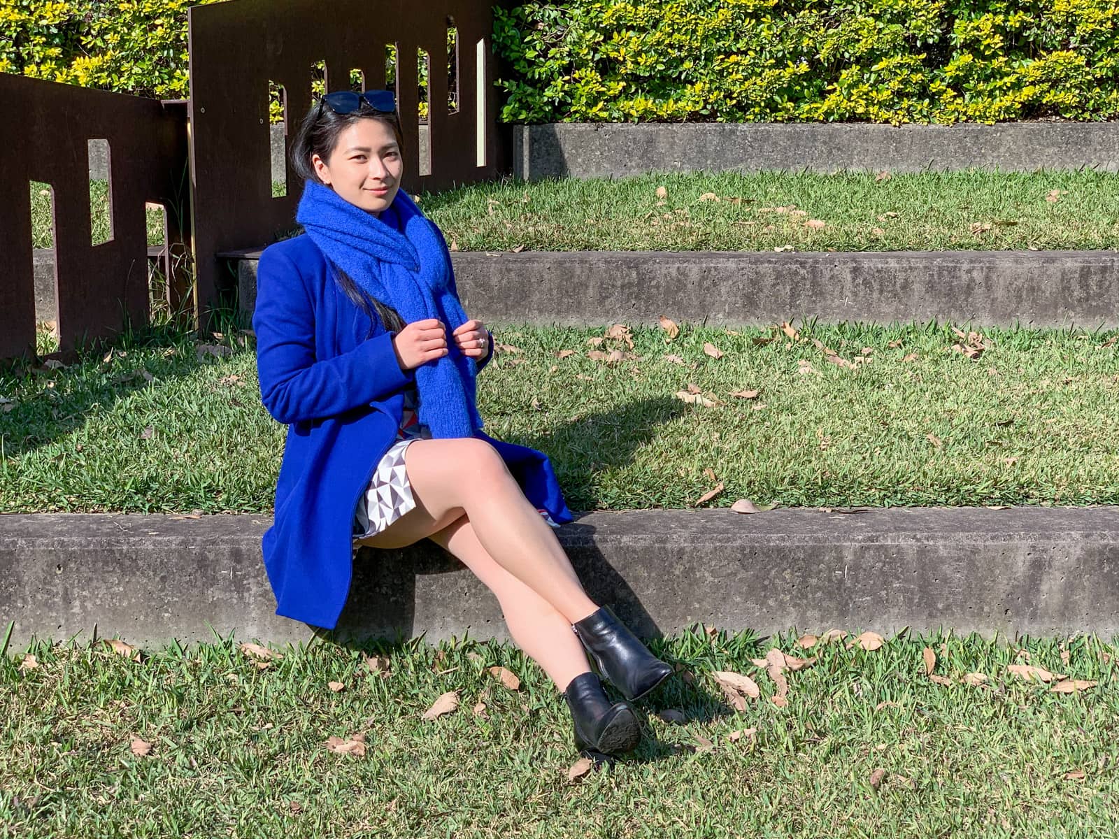 A woman wearing a bright blue coat and blue scarf, sitting on a concrete step in a grassy park. She is clutching the ends of her scarf and her legs are crossed slightly