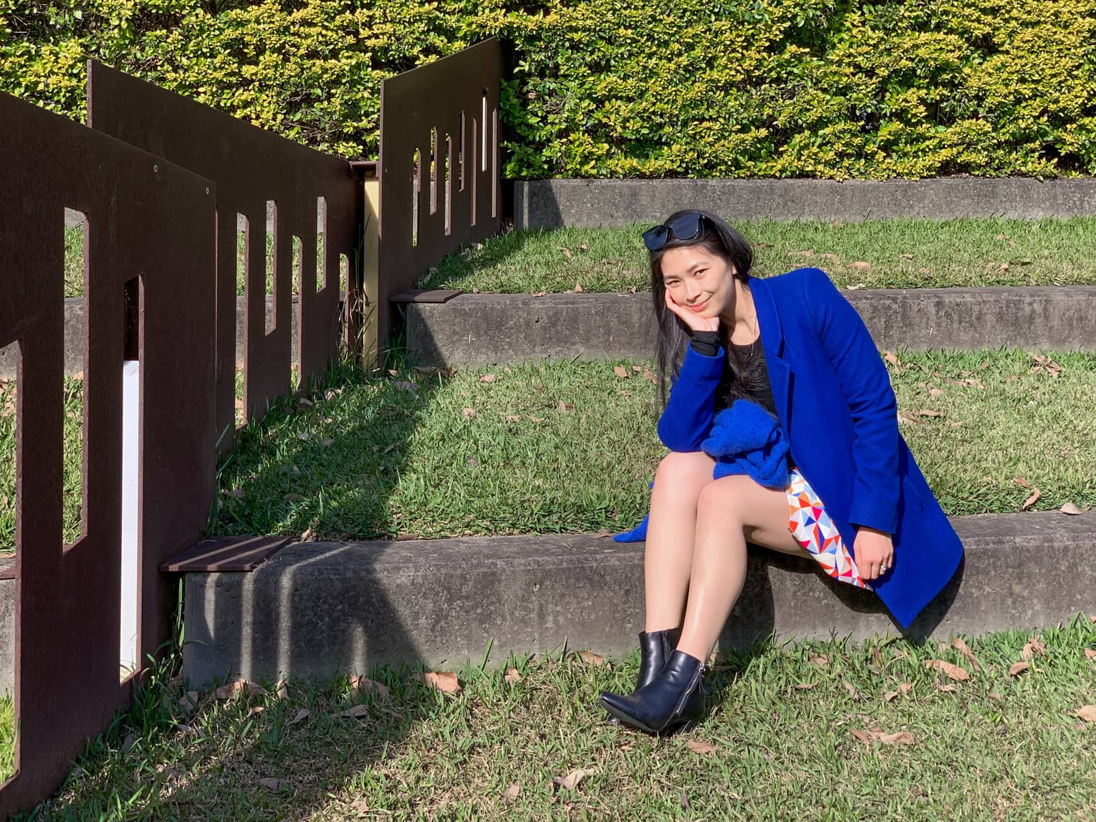 A woman with long dark hair sitting on a stone step in a grassy park. She has her knees bent and her head resting on her hand as her elbow is resting on her knee. She is wearing sunglasses on top of her head, and a bright blue coat. She is wearing a skirt and black heeled boots.