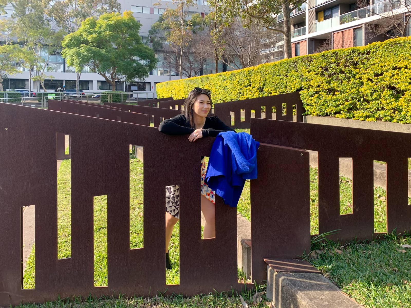 A woman in a black top leaning on a brown fence made of angular-cut, flat metal. She has a bright blue coat and scarf placed on part of the fence