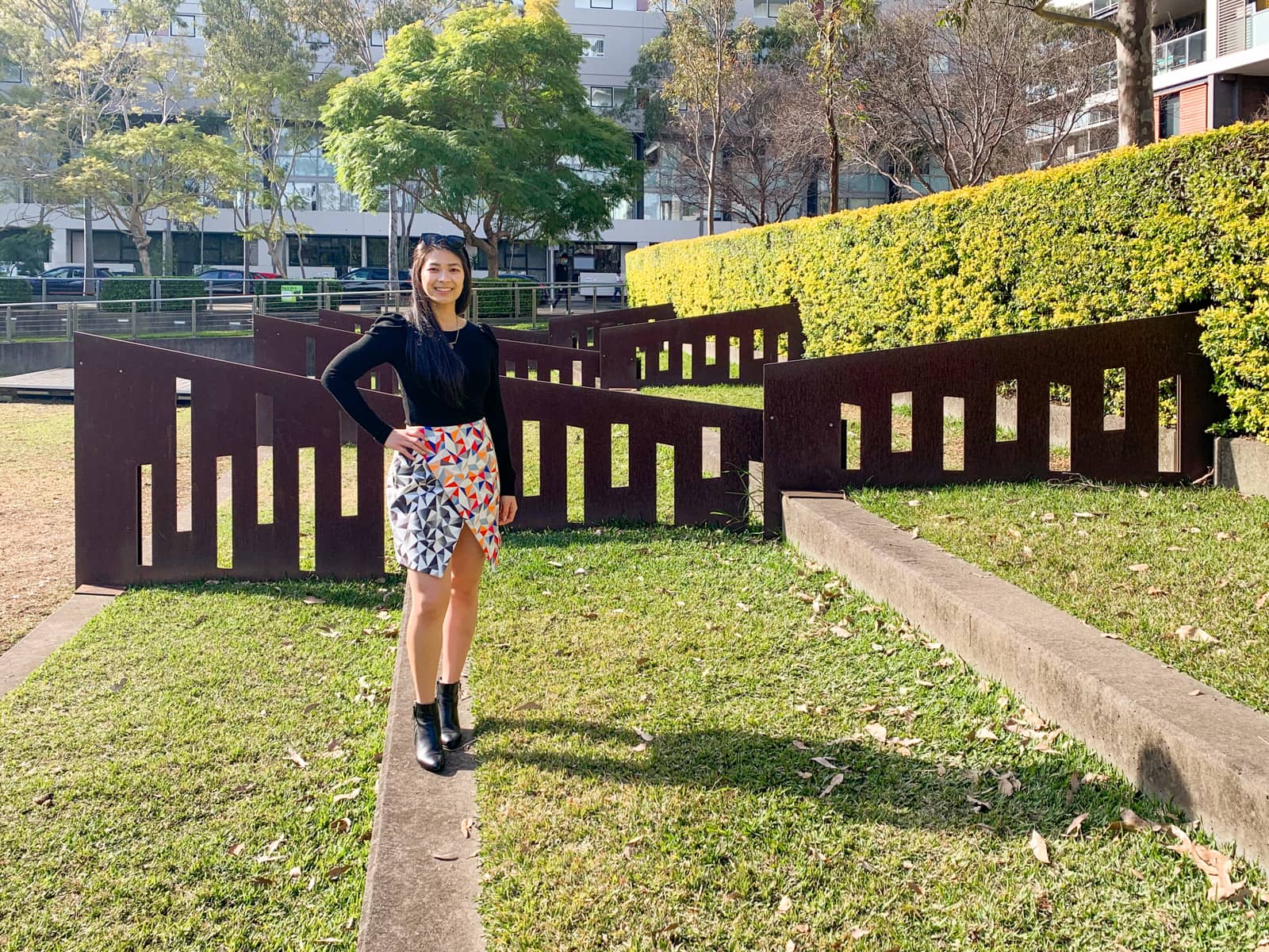 A woman standing in a park that has some interesting flat iron fencing. The woman is wearing a black top and black boots with a short, colourful geometric print skirt. She has a hand on her hip.