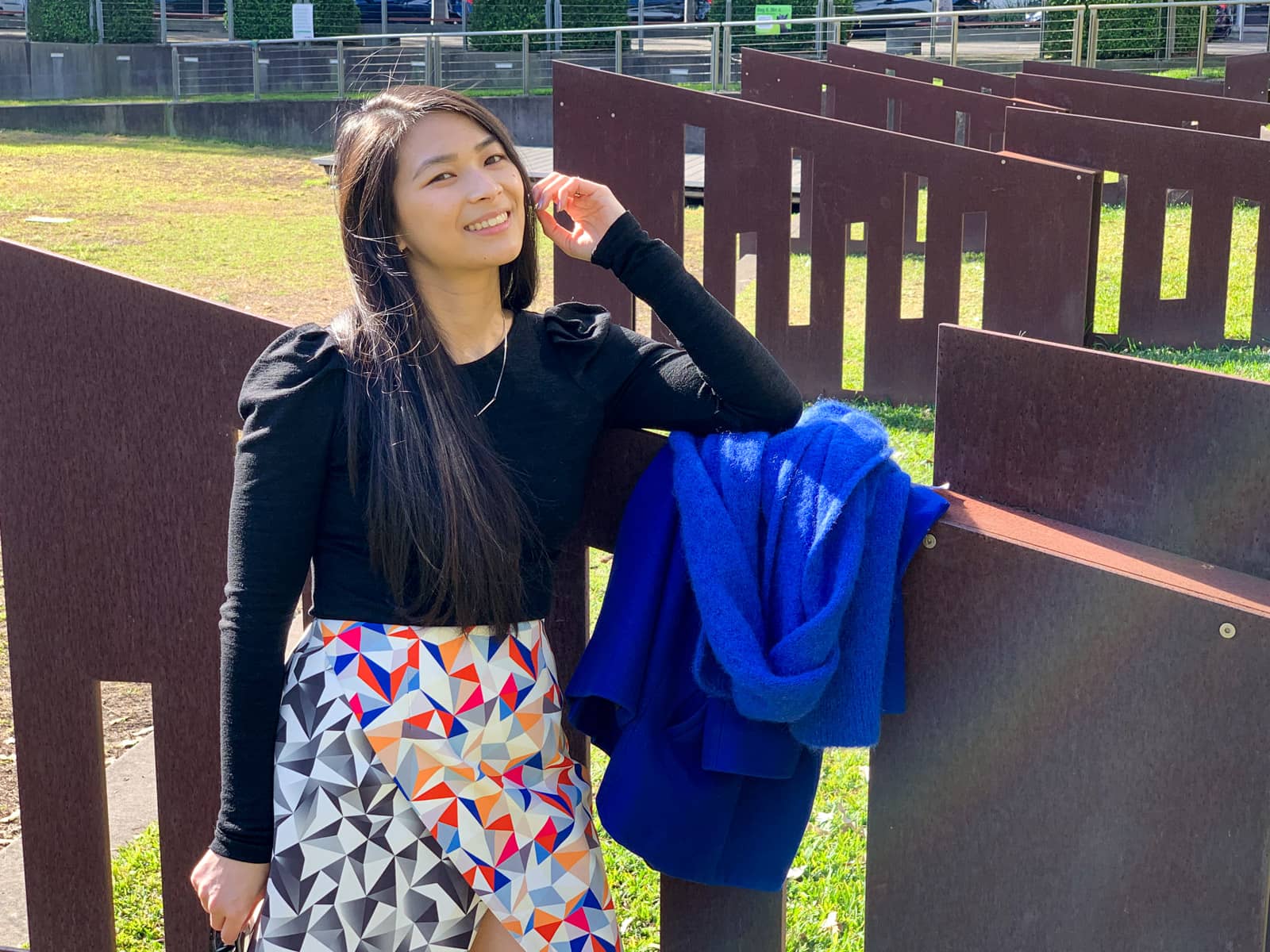 A close-up of a woman standing in a park leaning her elbow on a brown flat iron fence cut with rectangular holes. She is wearing a black top with puffy shoulders and has a blue coat and scarf on the fence next to her