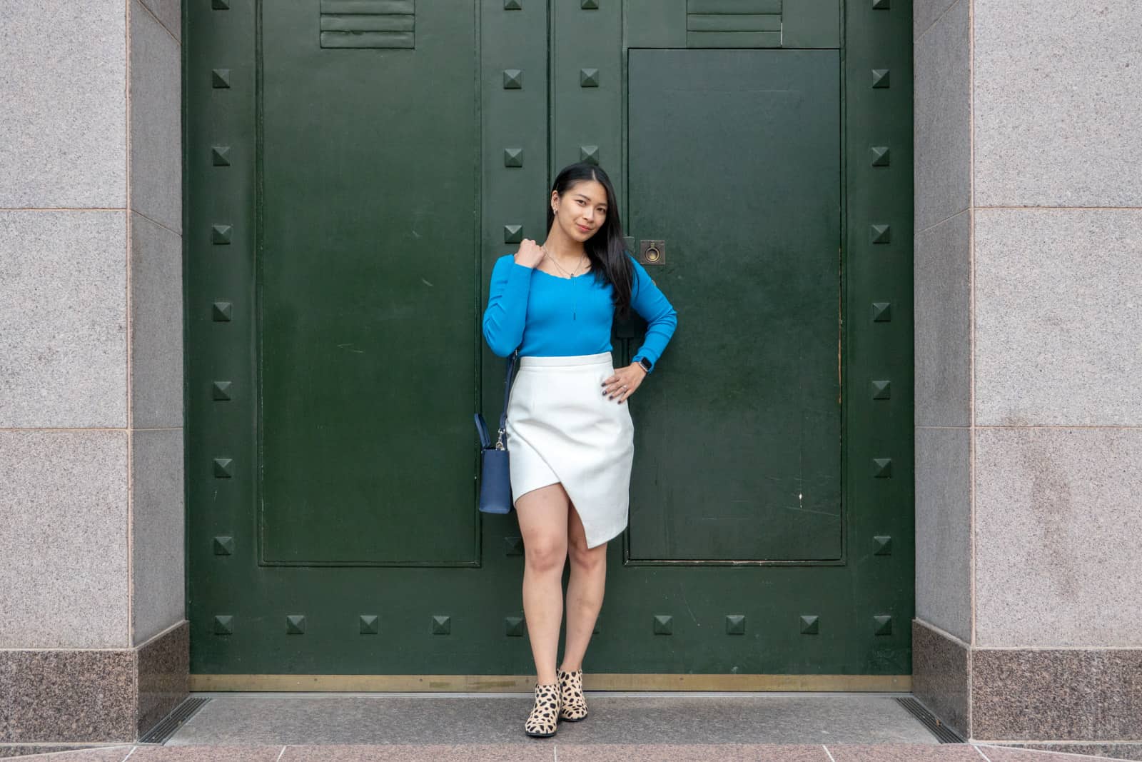 A woman standing in front of a very large dark green set of doors. She has short dark hair and is wearing a bright light blue long-sleeved top, a white wrap skirt, and shoes with animal print. She is carrying a small navy handbag on her shoulder and has a hand on her hip.