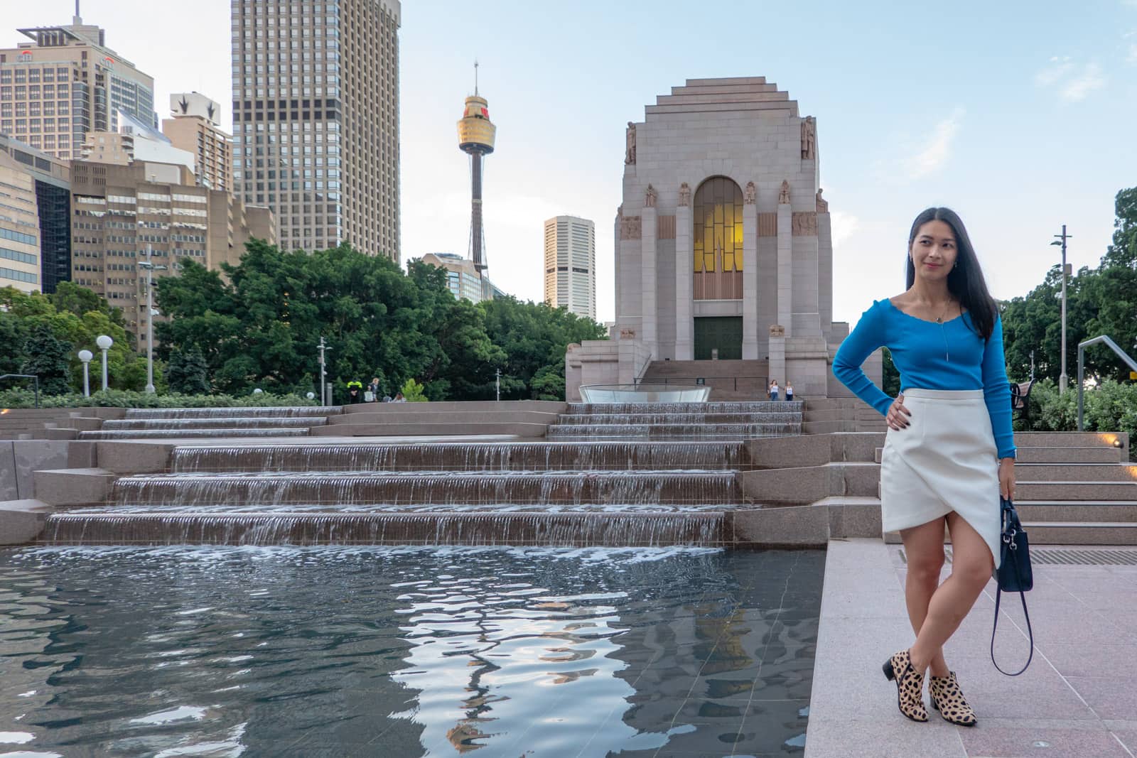 The same woman in other photos on this page, in the same outfit. She is standing by a reflecting pool with a water feature where the water flows down some steps. The city can be seen in the background.