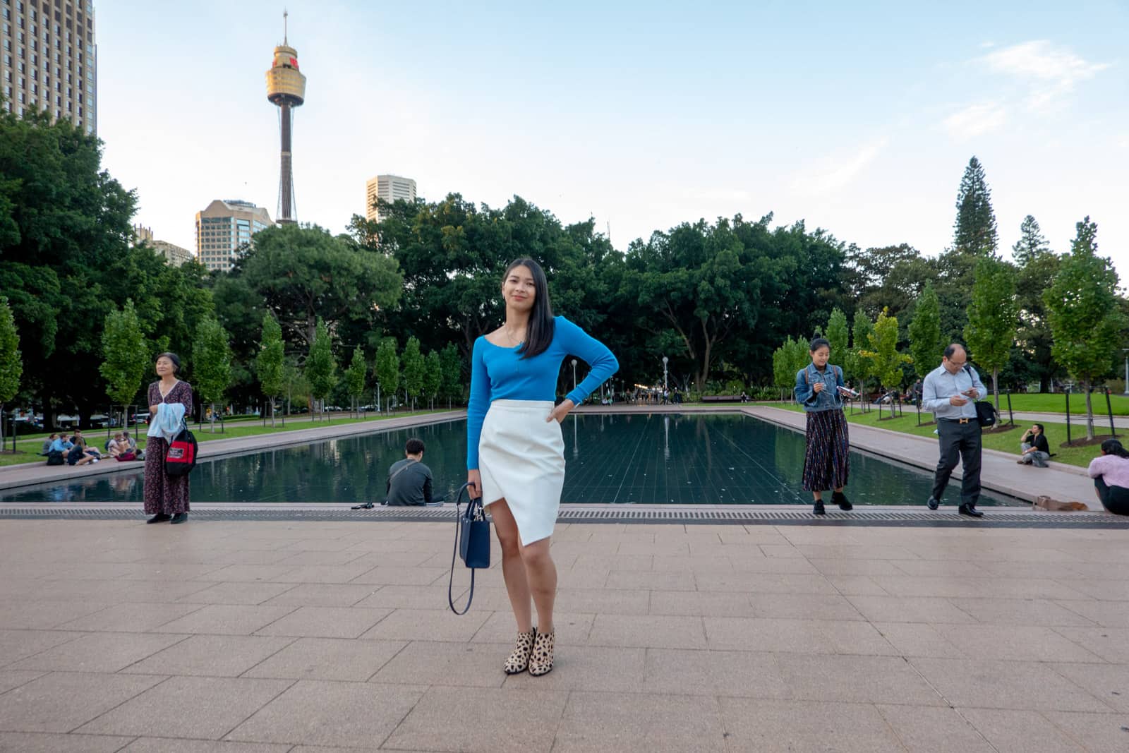 A view inside a park, with a rectangular water feature with still water. Centre of frame is the same woman in other photos on this page in the same outfit. There are a few other people minding their own business and standing or sitting near the water feature.