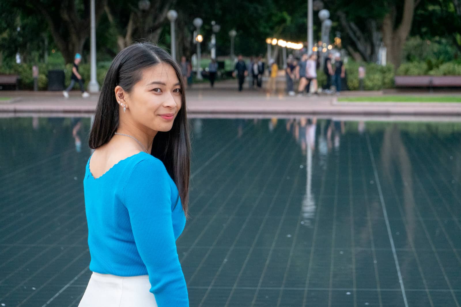 The same woman in other photos on this page, with her back slightly to the camera as she turns around and smiles and looks beyond the camera. In the background is a water feature with still water, that appears to be in a park.