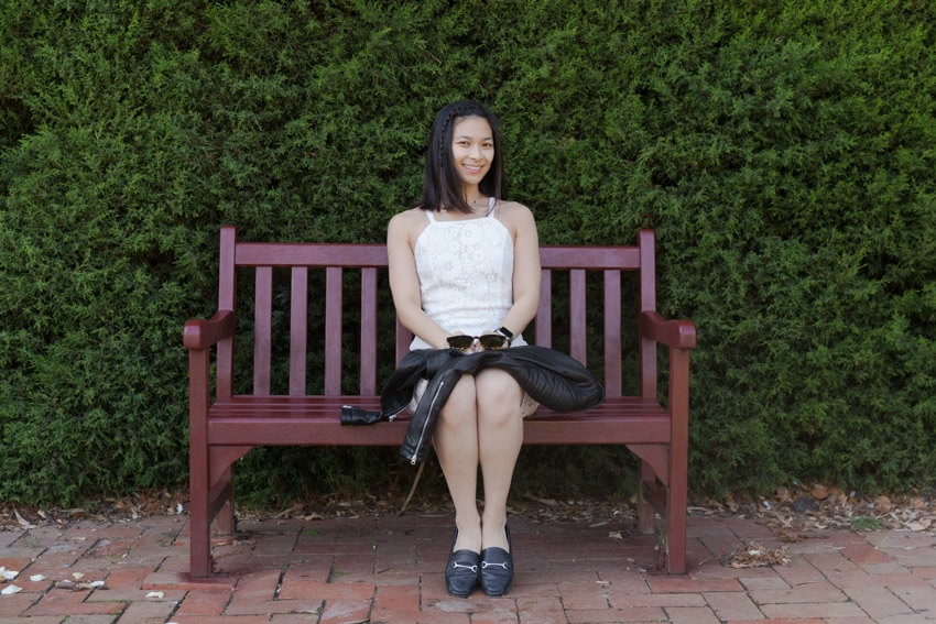 A woman in a white dress sitting on a dark red bench with a leather jacket on her lap. The background is a large hedge behind her.