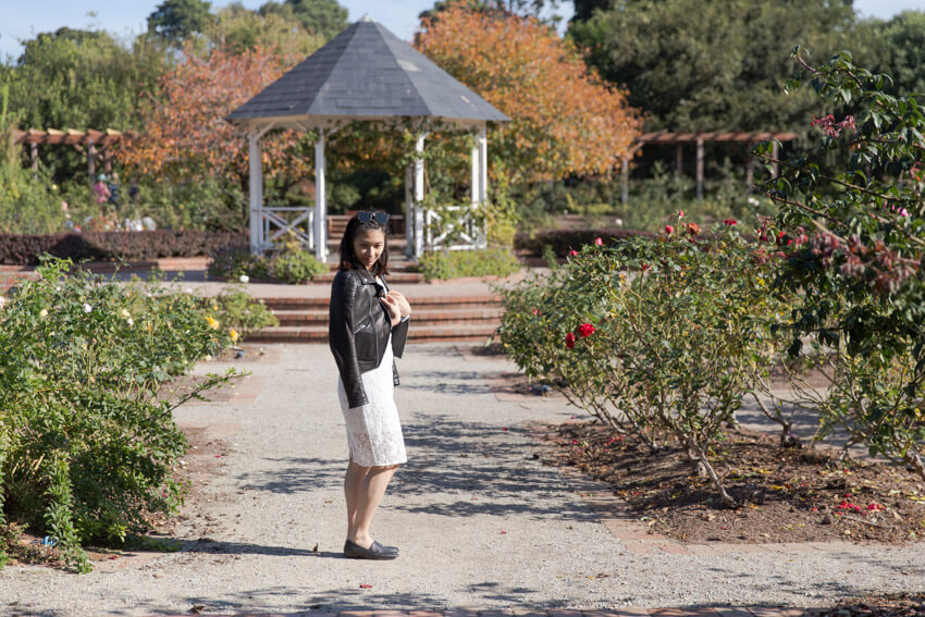 Side view of a woman wearing a white lace dress with a black leather jacket draped over her shoulders. She is standing in a rose garden with a dirt path and in the background is a gazebo.