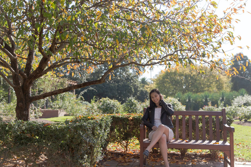 A woman with short dark hair sitting on a park bench. She is wearing a white lacy dress and a black leather jacket. The branches and leaves of the tree next to her frame the area above her head.