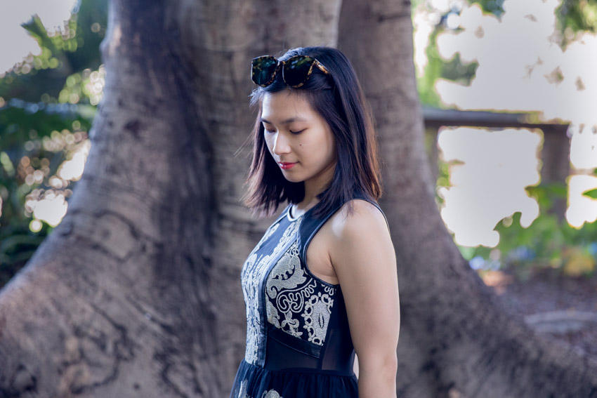 A woman with short hair, her eyes downcast. She is wearing a sleeveless black dress and standing side-on. She has tortoiseshell style sunglasses on top of her head.