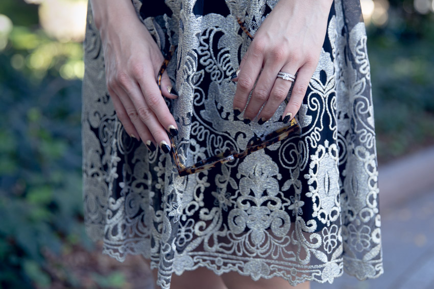 A close-up of a woman’s hands hanging in front of her black dress with gold embroidery. Her nails are black with glittery gold accents. She has a silver wedding and engagement ring on her left ring finger, and is holding onto a pair of tortoiseshell patterened sunglasses.