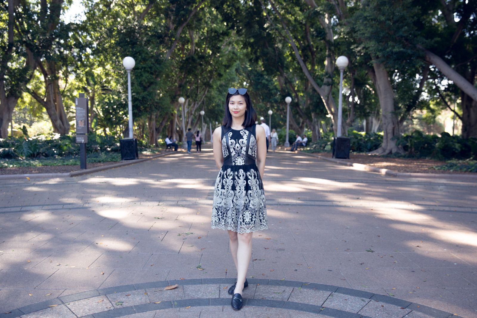 A woman wearing a black and gold sleeveless, knee-length dress. She has short dark hair and is wearing sunglasses on top of her head. She has her hands behind her back and is wearing black loafers. She is standing at the entrance to a park with a wide path extending behind her, lampposts on the edges of the wide paths and many tall trees in the background